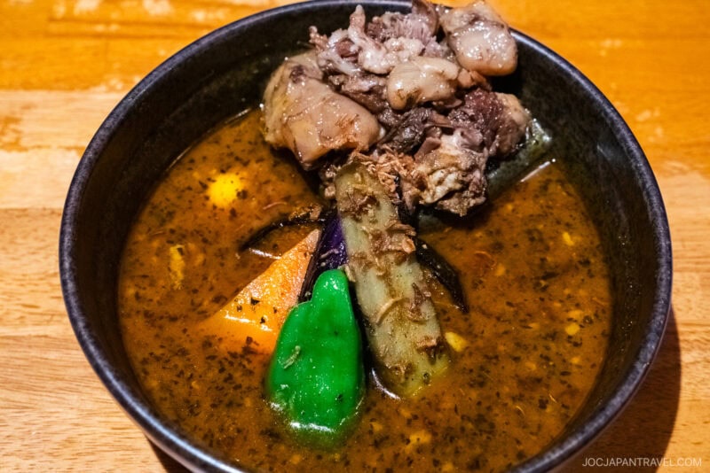 A bowl of Sapporo Soup Curry featuring tender pieces of meat, carrot, eggplant, green pepper, and other vegetables in a rich, dark broth, served in a black bowl on a wooden table.