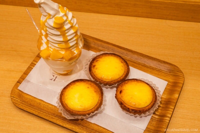 A wooden tray with three cheese tarts and a cup of vanilla soft serve ice cream topped with mango sauce and chunks, featured in the Sapporo Sweets Guide, set on a light wood table.