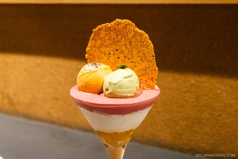 A parfait glass filled with layered mousse and topped with two ice cream scoops, a thin crispy tuile, and herbs&mdash;this elegant treat could grace the pages of any Sapporo Sweets Guide, set against a neutral brown background.