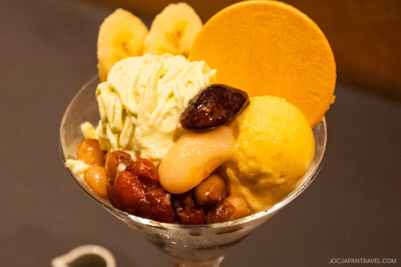 A parfait glass filled with green tea ice cream, yellow sorbet, sweet beans, banana slices, chestnut, white mochi, and a large round wafer—this Japanese dessert is a highlight in the Sapporo Sweets Guide.