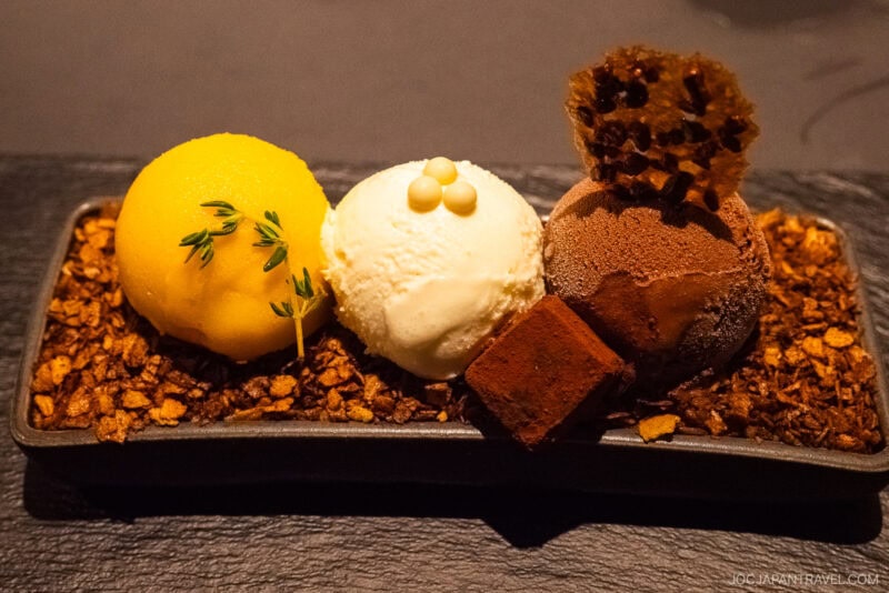 Featured in the Sapporo Sweets Guide, three scoops of gourmet ice cream—mango, vanilla, and chocolate—rest on crunchy granola, garnished with a chocolate cube, honeycomb, and thyme on a sleek black dish.