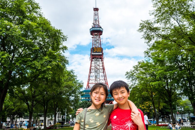 Two smiling children stand arm-in-arm in front of the iconic Sapporo TV Tower in Japan, surrounded by green trees under a partly cloudy sky. The digital clock on the Sapporo TV Tower clearly displays 3:00.