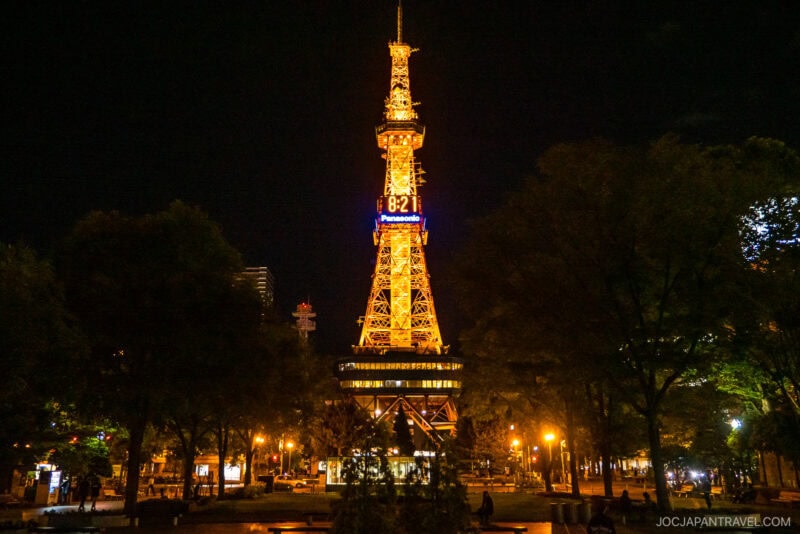 The Sapporo TV Tower illuminated at night, glowing with yellow lights and displaying the time 8:21 at the top. Trees and street lamps add charm to this stunning view of the iconic Sapporo TV Tower.