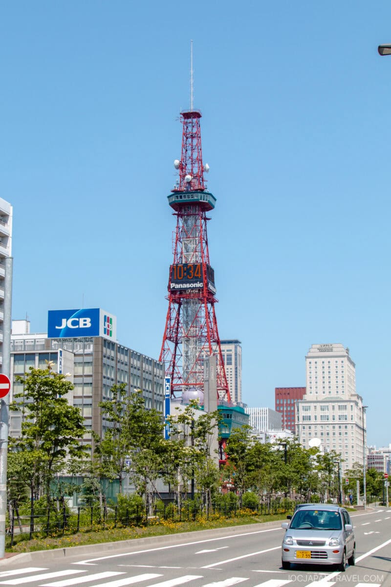 A red and white Sapporo TV Tower with a digital clock stands among modern city buildings under a clear blue sky; cars and greenery line the street in the foreground.