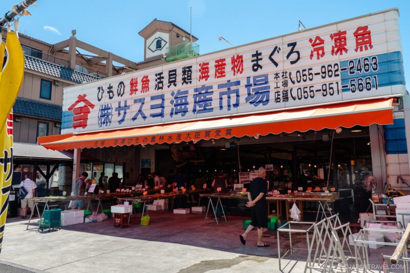 Outdoor seafood market in Numazu, Japan, with a large sign in Japanese above the entrance. People shop for seafood under a covered area filled with tables of fresh products. Bright daylight and a blue sky overhead.