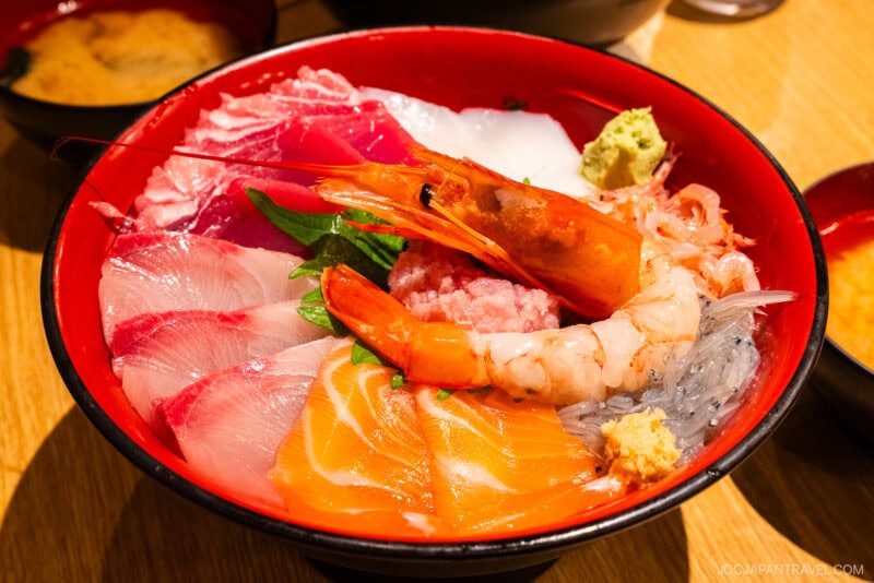 A colorful bowl of assorted sashimi, featuring shrimp, salmon, tuna, and other raw fish slices served on rice and garnished with wasabi and ginger&mdash;a true Numazu specialty. Miso soup is in the background.