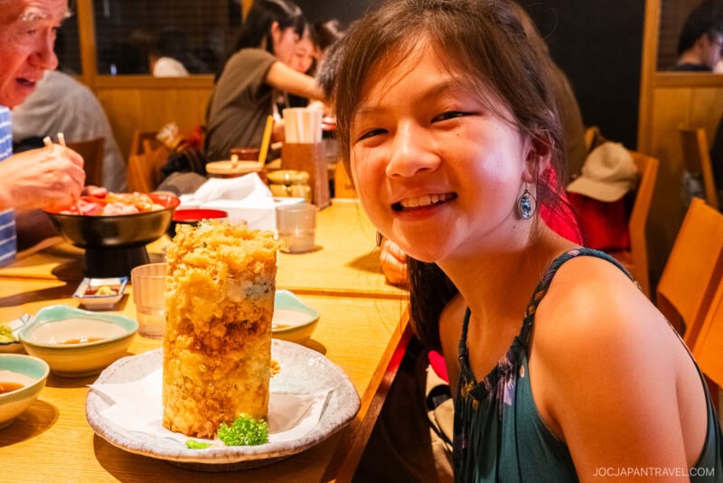 A smiling girl sits at a restaurant table in Numazu, with a tall, cylindrical tower of tempura in front of her, surrounded by small dishes and other diners in the background.