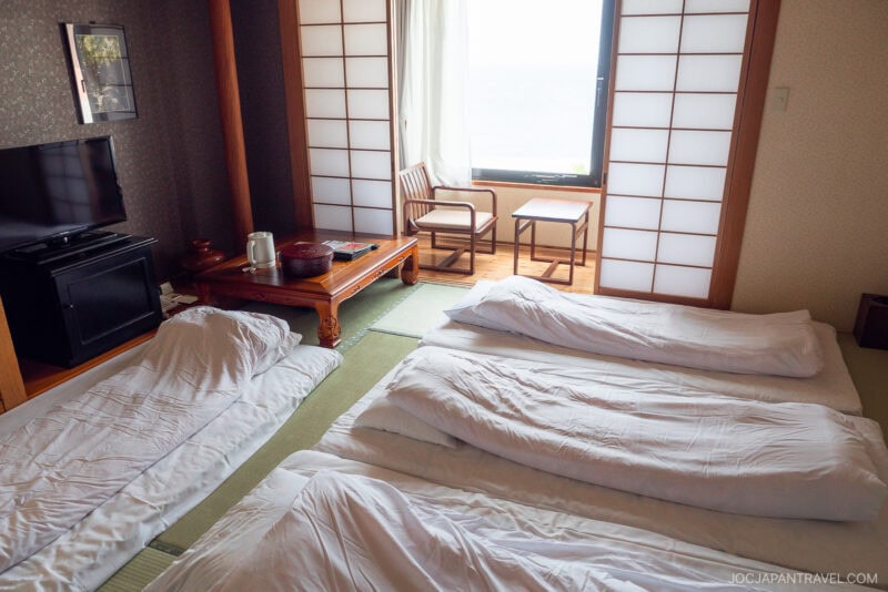 A traditional Japanese tatami room at Seaside Hotel Yakushima with three futon beds on the floor, a low wooden table with cushions, a TV, shoji screen doors, and a window overlooking a bright outdoor view.