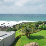 View of a grassy lawn with palm trees at Seaside Hotel Yakushima, bordered by modern buildings, overlooking rocky shorelines and the ocean under a cloudy sky. Waves crash against the rocks in the distance.