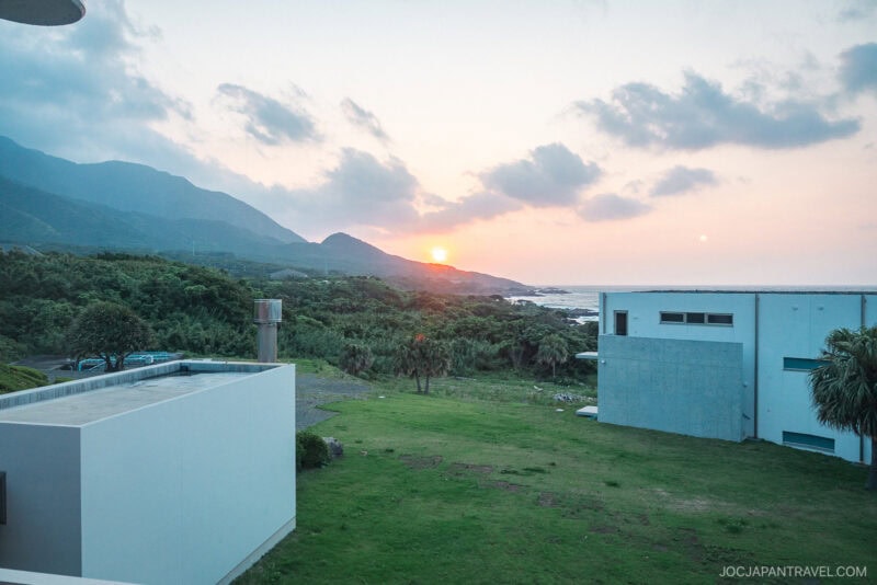 View of a grassy yard bordered by modern white buildings at Seaside Hotel Yakushima, with mountains and lush greenery in the background and the sun setting over the ocean under a partly cloudy sky.