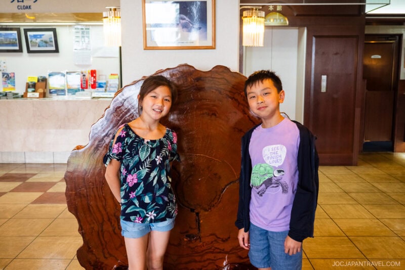 Two children stand smiling in front of a large, polished tree cross-section display in the Seaside Hotel Yakushima lobby, with the reception and elevator visible in the background.