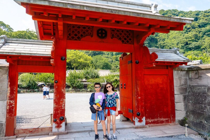 Two people stand smiling under a large traditional red Japanese gate at Sengan-en, with greenery, stone walls, and a bright sky in the background. They are wearing sunglasses and holding fans.