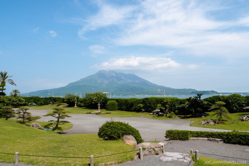 A lush Japanese garden at Sengan-en with manicured trees and stone paths overlooks the ocean, framed by a large volcanic mountain and a partly cloudy sky in the background.