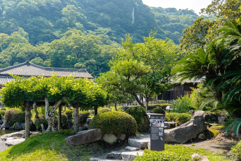 A lush Japanese garden at Sengan-en features trimmed bushes, trees, large rocks, and a small stream. Traditional wooden buildings and forested hills rise in the background under a sunny sky.