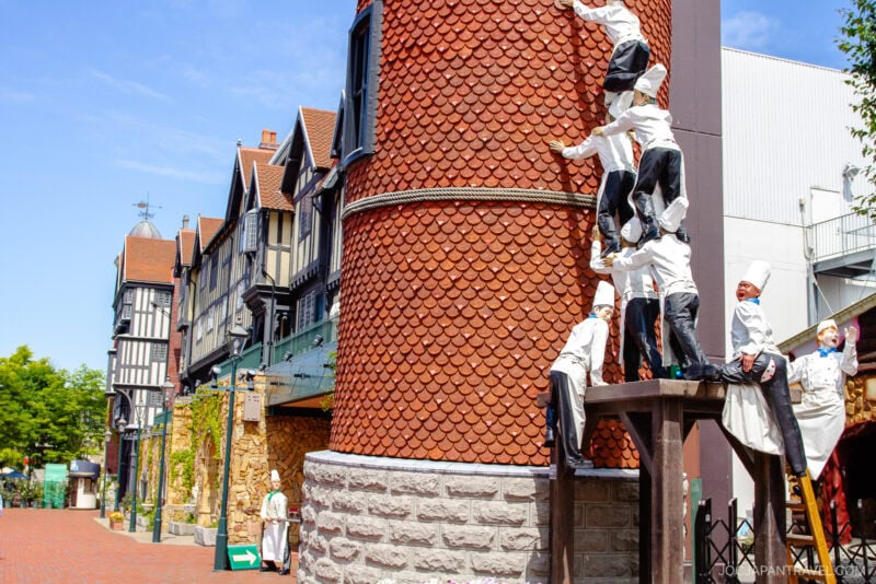 A whimsical outdoor scene in Shiroi Koibito Park features statues of chefs climbing up a red-brick tower in a European-style village. Tudor-style buildings line the sunny street in the background, adding to the park's fairytale charm.