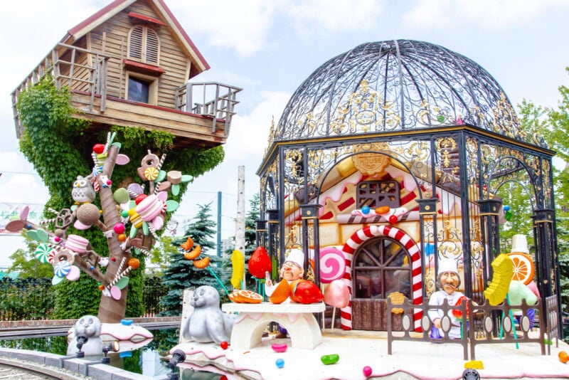A whimsical scene at Shiroi Koibito Park features a candy-themed house with a dome roof, colorful treats, chefs, playful animal statues, and a treehouse surrounded by lush greenery and a bright sky.
