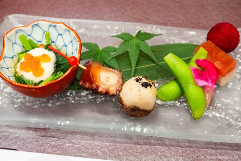 A beautifully arranged Japanese appetizer platter with assorted items, served on a glass tray with a green leaf underneath—just one of the elegant culinary experiences at Shiroyama Hotel 城山ホテル鹿児島: A Room with a View in Kagoshima’s Hilltop Retreat.
