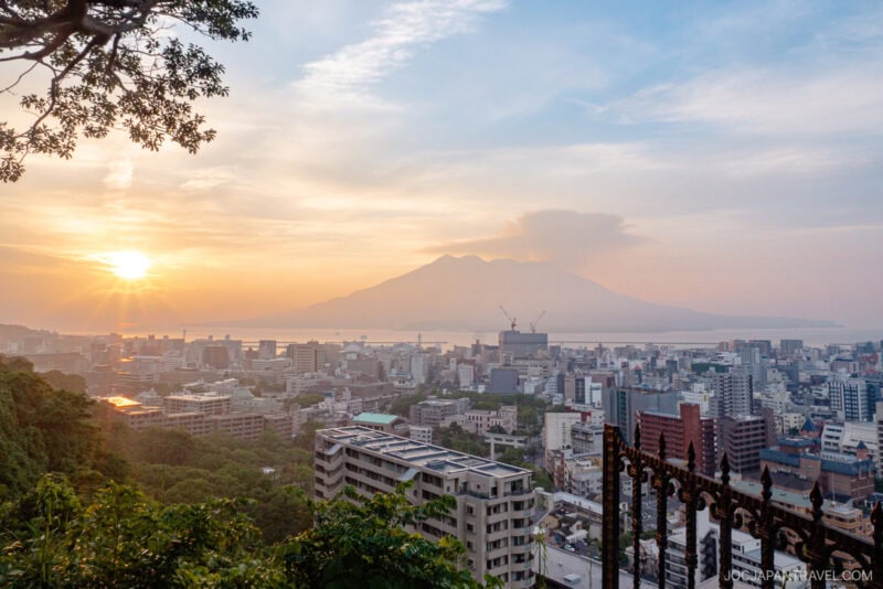 Sunrise over a cityscape with a distant volcano, partially veiled by clouds, rising behind the buildings&mdash;just the view from Shiroyama Hotel 城山ホテル鹿児島, Kagoshima&rsquo;s hilltop retreat. Trees frame the scene as warm light bathes the peaceful morning.