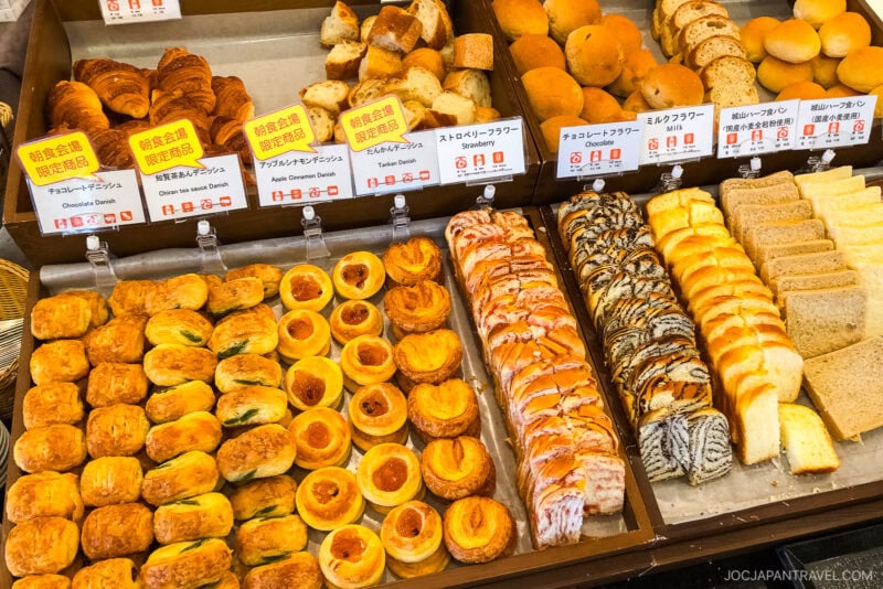 Assorted pastries and breads displayed in a bakery at Shiroyama Hotel 城山ホテル鹿児島: A Room with a View in Kagoshima’s Hilltop Retreat, with signs in Japanese and English indicating flavors and ingredients.