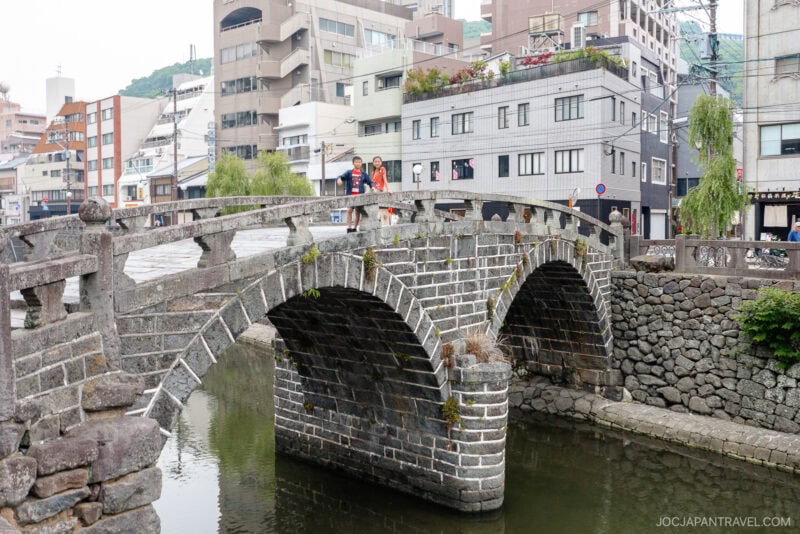 Stone arched bridge over the Nakashima River canal in a city, with two people standing on the bridge; modern buildings and greenery visible in the background.