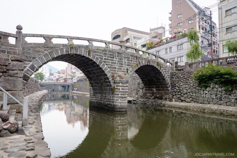 A historic stone arch bridge crosses the calm Nakashima River, surrounded by stone walls and urban buildings. Trees and greenery line the waterway, creating a peaceful and scenic atmosphere.