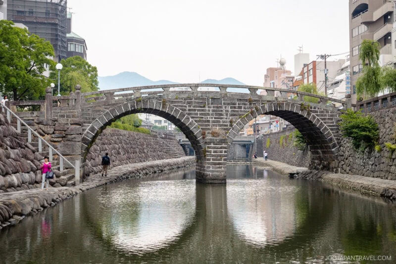 Two stone arch bridges span the calm Nakashima River in an urban area, with people strolling along stone-paved walkways; buildings and trees line the scenic riverbanks.