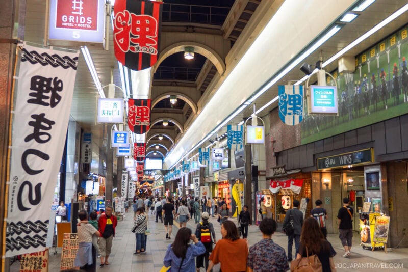 A busy covered shopping street in Japan, Tanukikoji Shopping Street features colorful banners, various shop signs, and many people walking and shopping under bright lights.