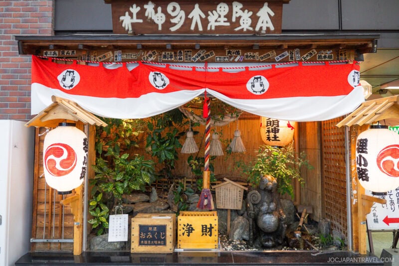 A small Shinto shrine along Tanukikoji Shopping Street features red and white lanterns, wooden signs, and a stone tanuki statue, all decorated with greenery and a sacred rope under a wooden roof with Japanese writing.