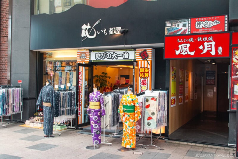 A kimono shop along Tanukikoji Shopping Street in Japan displays colorful kimonos and garments on mannequins outside its entrance. The shop has a black and orange sign with Japanese text, beside a red restaurant adorned with hanging lanterns.