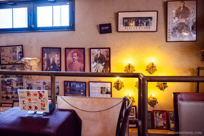 A cozy cafe interior with framed photos, posters, and memorabilia on the walls, soft yellow lighting from wall sconces, and a table set with menus featuring Toruko Rice in the foreground.