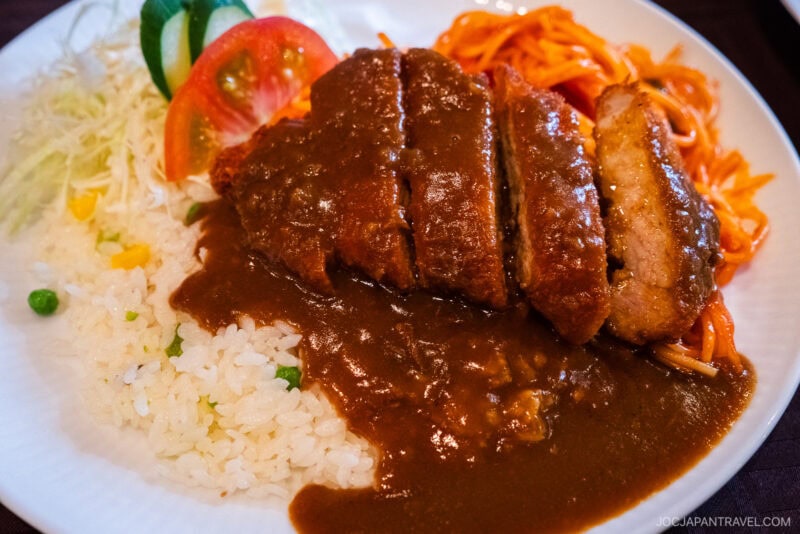 A plate of Toruko Rice featuring Japanese curry rice topped with breaded fried pork cutlet, shredded cabbage, tomato, cucumber, and pasta, all served with a rich brown curry sauce.