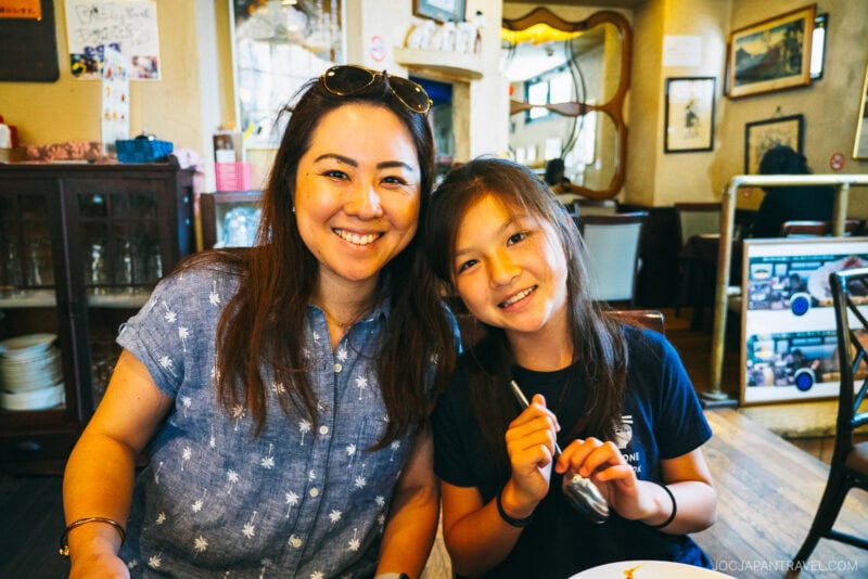 A woman and a young girl smile while sitting together at a cozy, warmly lit restaurant. The girl holds a fork, ready to enjoy Toruko Rice, and both appear happy and relaxed amid tables, chairs, and charming decor.