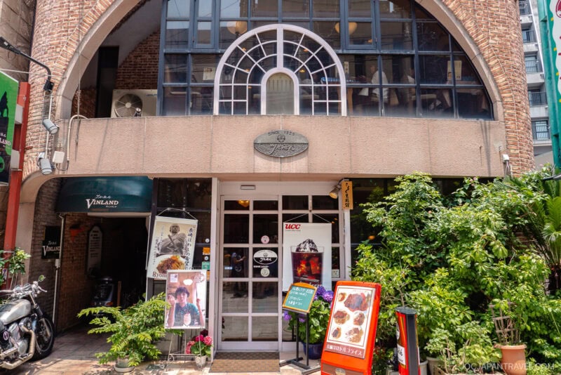A café entrance with large windows, plants, and colorful signs displaying food and drink options like Toruko Rice. Posters and decorations adorn the door and walls, while arched brickwork frames the building’s welcoming facade.