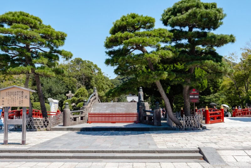 A traditional Japanese stone bridge with red railings at Tsurugaoka Hachimangu is framed by tall green pine trees under a clear blue sky. Stone pavement leads up to the bridge, and several visitors are nearby.