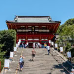 People walk up a wide stone staircase leading to the Tsurugaoka Hachimangu shrine, with its red and white facade and green-tiled roof, surrounded by trees under a clear blue sky.