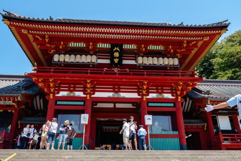 Visitors stand on steps and gather in front of Tsurugaoka Hachimangu, a large traditional Japanese shrine with red pillars, ornate details, and lanterns, set against a clear blue sky and trees.