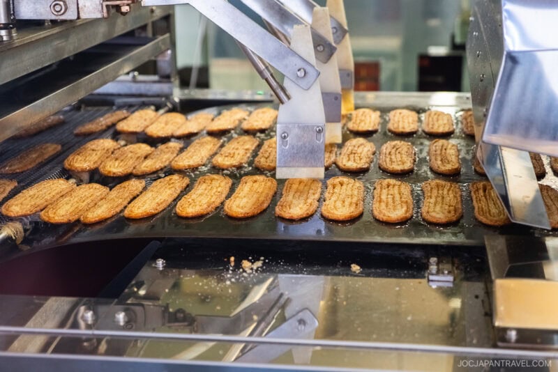 Rows of rectangular cookies move along a conveyor belt in the Unagi Pie Factory, passing under a metal mechanism during the production process.