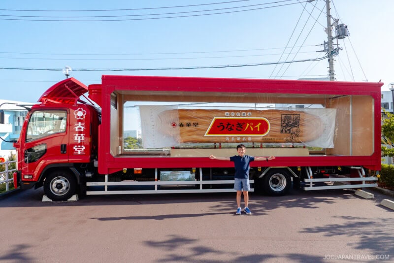A person stands with arms outstretched in front of a red truck from the Unagi Pie Factory, displaying a giant packaged pastry resembling unagi pie inside a large glass case mounted on the truck bed.