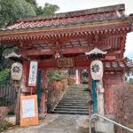 Yakuoin Onsenji Temple (薬王院温泉寺) – A Spiritual Stop in Yamashiro Onsen A traditional Japanese temple gate with a tiled roof, lanterns, and wooden pillars marks the entrance to Yakuoin Onsenji Temple. Stone steps lead upward through the gate, surrounded by trees and signs with Japanese writing under an overcast sky.