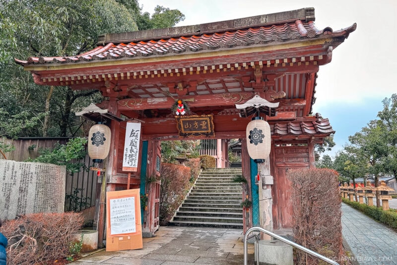 A traditional Japanese temple gate with a tiled roof, lanterns, and wooden pillars marks the entrance to Yakuoin Onsenji Temple. Stone steps lead upward through the gate, surrounded by trees and signs with Japanese writing under an overcast sky.
