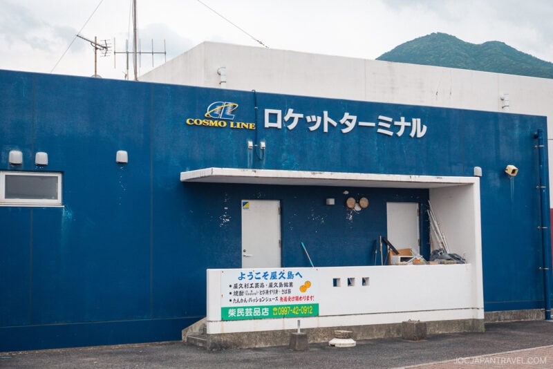 A blue building with a sign reading Cosmo Line Rocket Terminal in Japanese and English. A white board with information stands out front, while mountains loom in the background. The weathered terminal serves both Toppy & Rocket ferries.