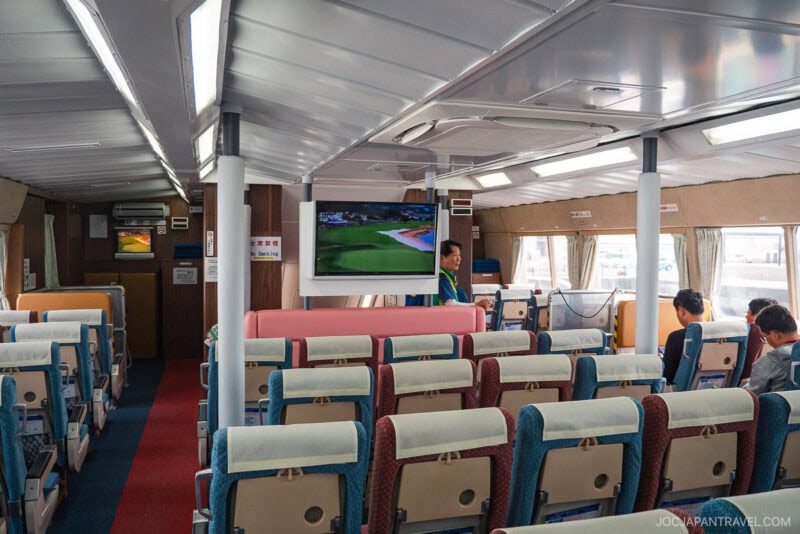 The interior of a ferry with rows of cushioned seats, passengers sitting, and a large screen at the front displaying a Toppy & Rocket golf game. Windows let in daylight, and a crew member stands near the screen.