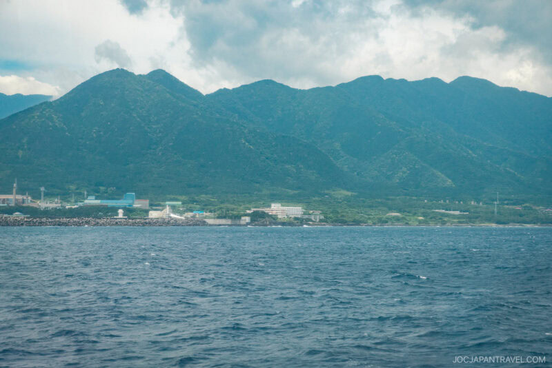 A view of a coastal town with Toppy buildings along the shore, lush green mountains in the background, and choppy Rocket-blue ocean in the foreground under a cloudy sky.