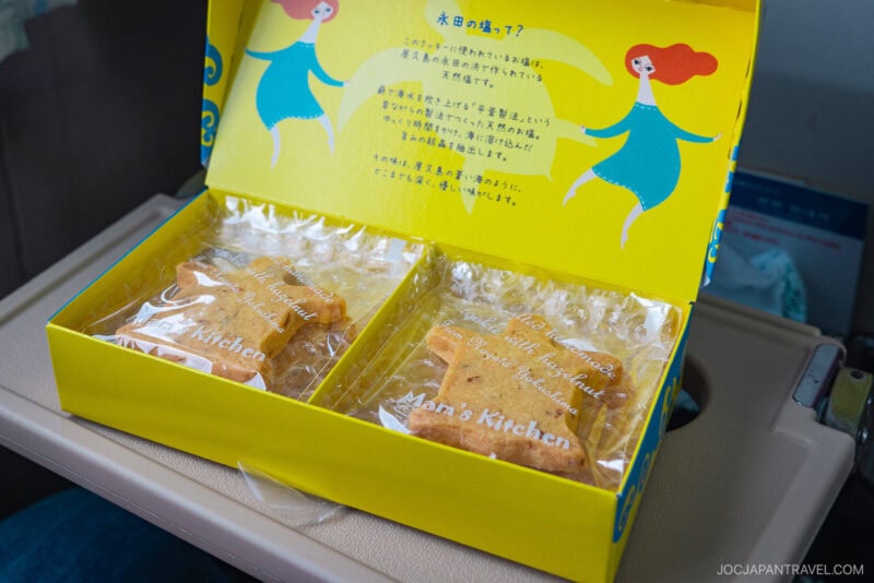 A yellow box with illustrations and Japanese text contains two types of individually wrapped baked goods, labeled Man’s Kitchen, featuring Toppy & Rocket, placed on an airplane tray table.