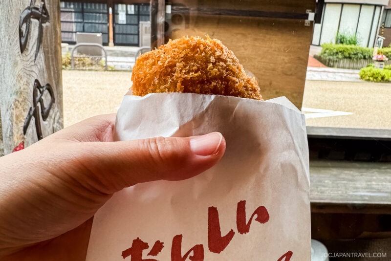 A hand holding a breaded and fried snack in a white paper wrapper with red lettering, outdoors near a wooden structure in Yamanaka Onsen, with a street visible in the background.