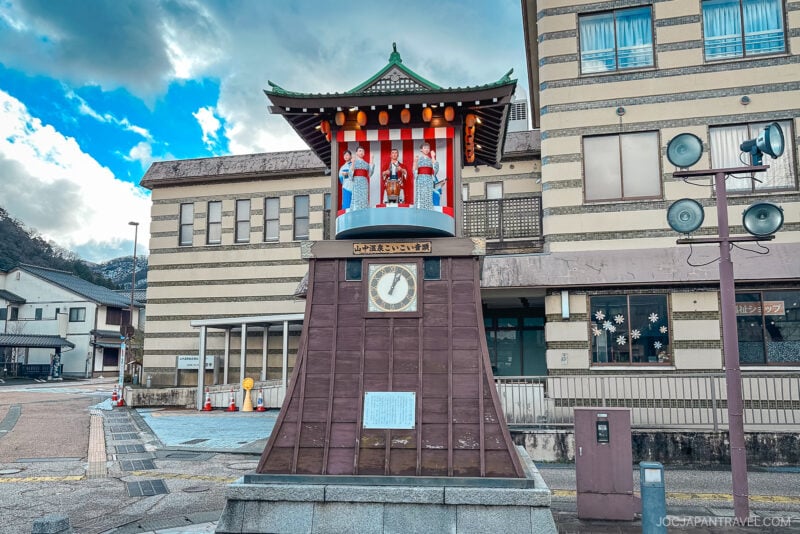 A traditional Japanese clock tower with mechanical dolls stands in Yamanaka Onsen, set against a striped red and white backdrop and a beige building, on a street beneath a cloudy sky.