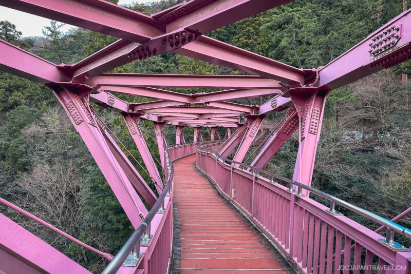 A winding pedestrian bridge with red decking and pink steel beams curves through a forested hillside near Yamanaka Onsen, surrounded by green and brown trees.