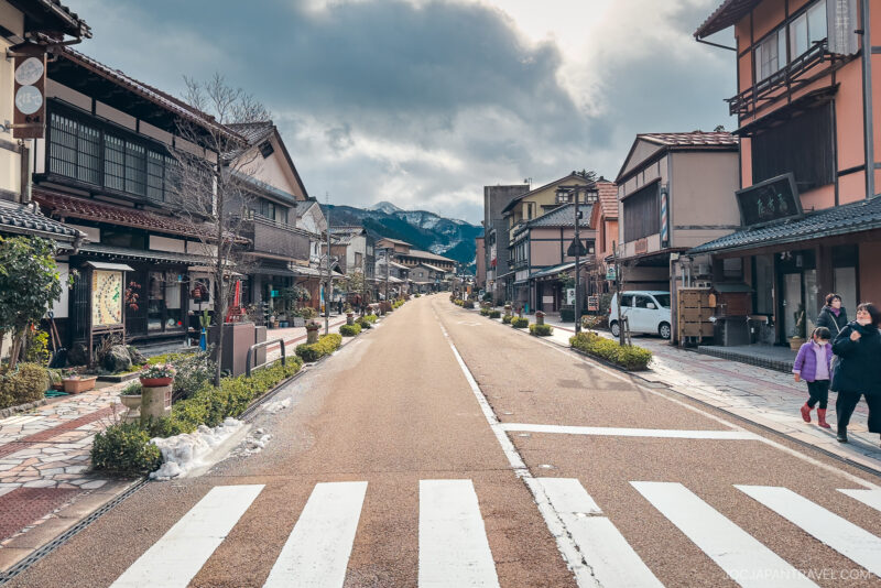 A quiet, empty street in the Japanese town of Yamanaka Onsen with traditional wooden buildings on both sides, mountains in the distance, and a few people walking on the sidewalk under a cloudy sky.
