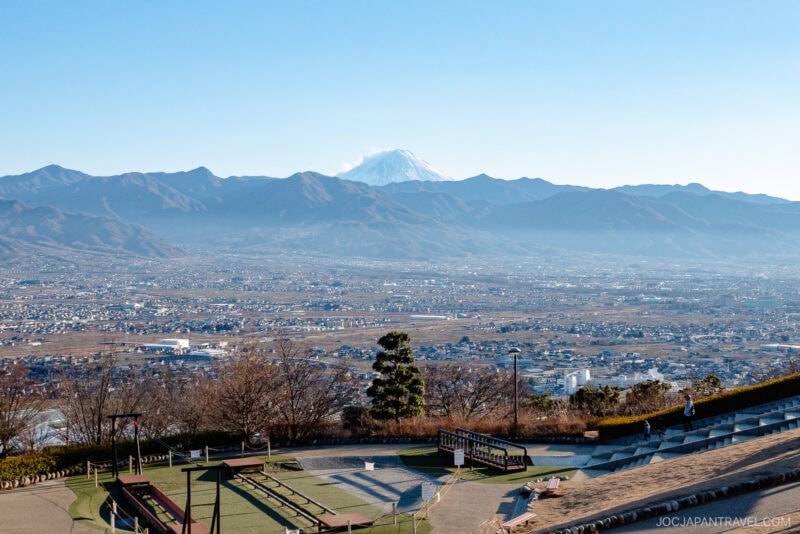 A scenic view of a wide valley in Yamanashi with a city below, surrounded by mountains. Mount Fuji, snow-capped and partly shrouded in clouds, rises in the distance. In the foreground, there is a park with benches and walkways.