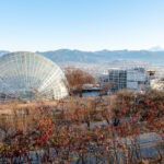 Yamanashi Fruit Picking and Wine Tasting – A Countryside Escape Two large glass-domed conservatories sit in a park in Yamanashi, with mountains and a city visible in the background under a clear blue sky. Bare trees and shrubs are in the foreground.